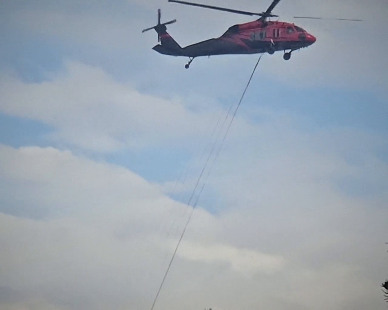 A red-and-black helicopter flying a longline against the clouds