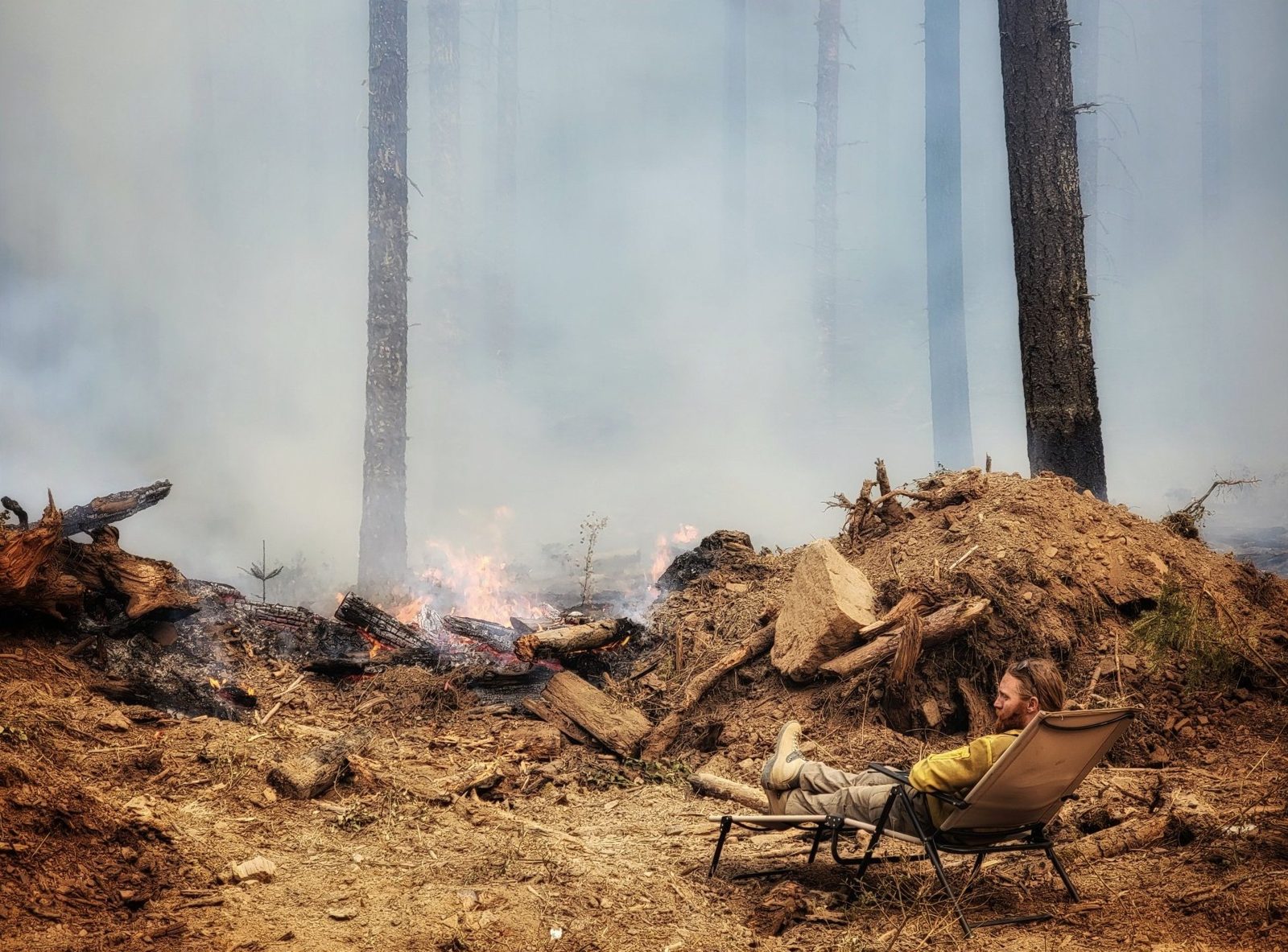 A camp chair beside a smoking stump pile in a burned forest