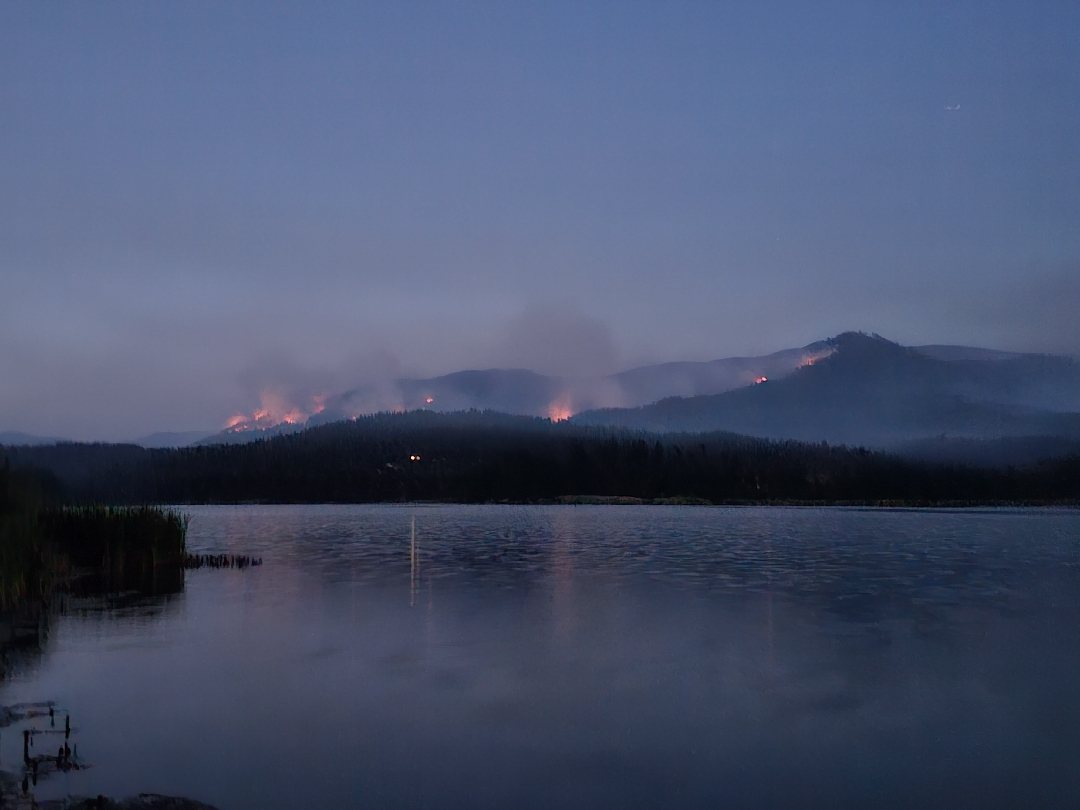 A wildfire burning on a ridge across a lake at dusk